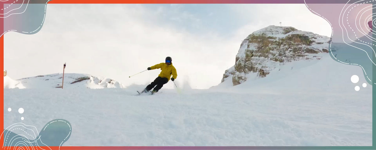 Skier in a bright yellow jacket carving through fresh snow on a mountain slope, surrounded by snow-covered rocky peaks and winter scenery.
