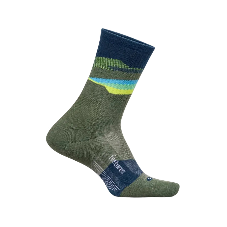 A single crew sock shown on a foot form against a light beige background. The sock is olive green with a mountain-like wave pattern in navy, green, and yellow near the cuff, and features the 'feetures' brand name on the heel.