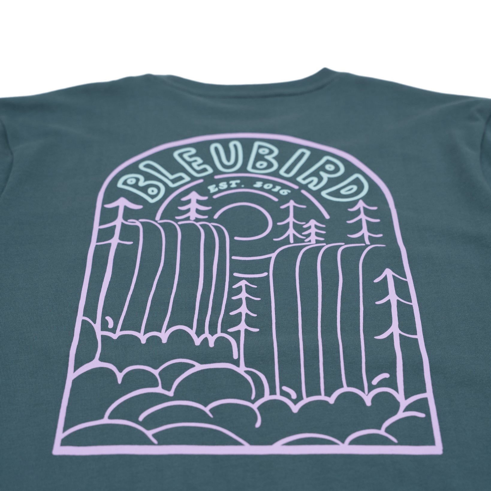 A close-up of the back of a dark teal-green t-shirt featuring a large light purple line art graphic of a waterfall landscape with the text 'BLEUBIRD EST. 2016'.