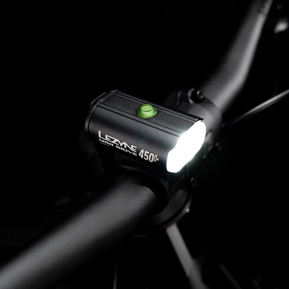 A black Lezyne light bar 450+ bicycle headlight mounted on a dark bicycle handlebar, with the front light illuminated and a small green button on top.